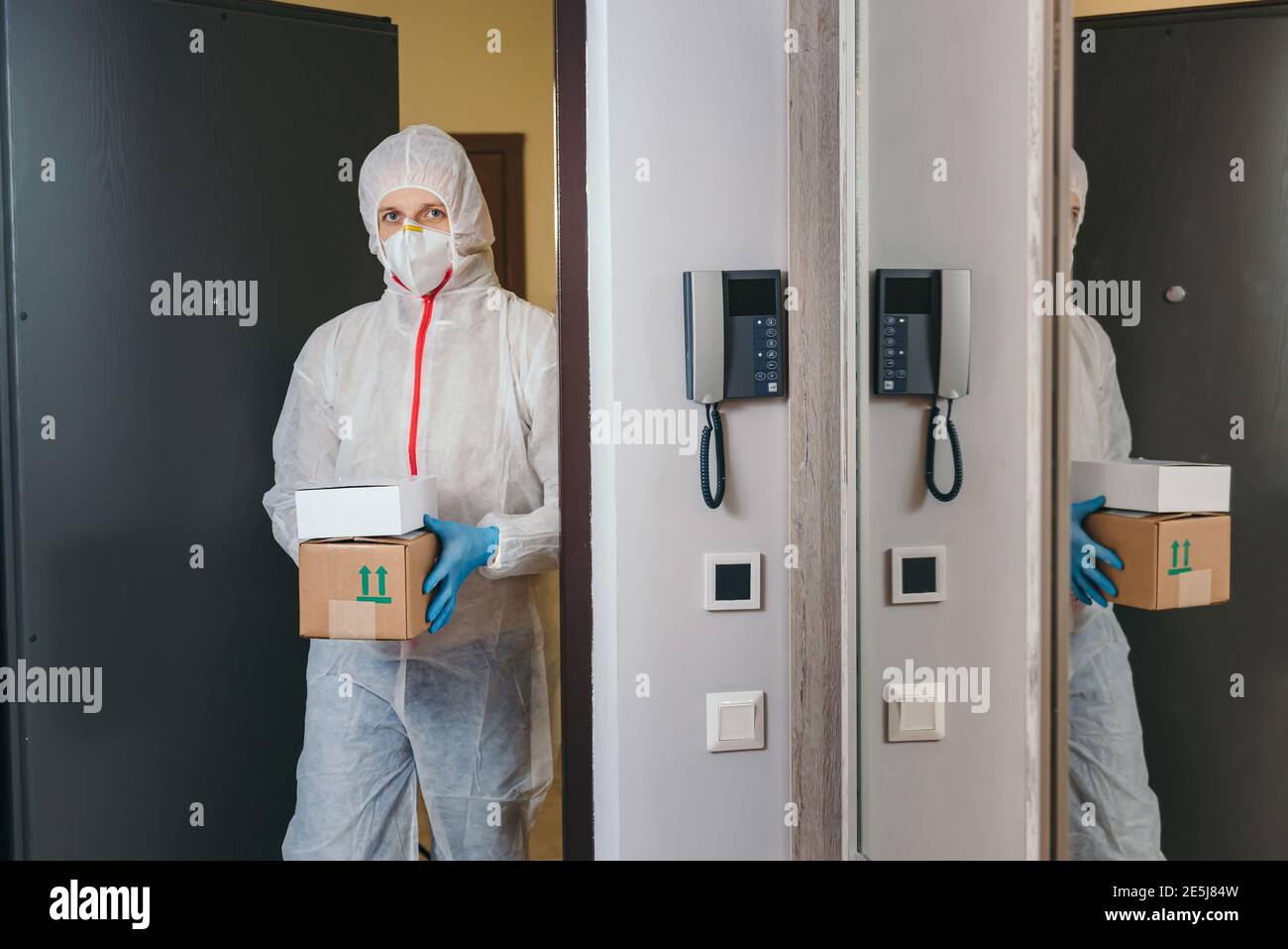 Mann hält Papierkiste mit Waren in medizinischen Handschuhen und Schutzmaske, kopieren Raum. Sichere Lieferung per Kurier während Coronavirus Quarantäne. Stockfoto