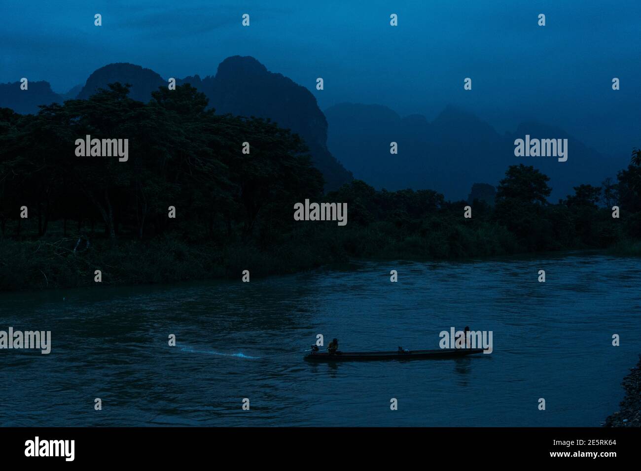 Zwei Männer in einem kleinen Boot, die in der Abenddämmerung in Vang Vieng, Laos, einen Fluss überquerten, mit Bergen im Hintergrund. Stockfoto