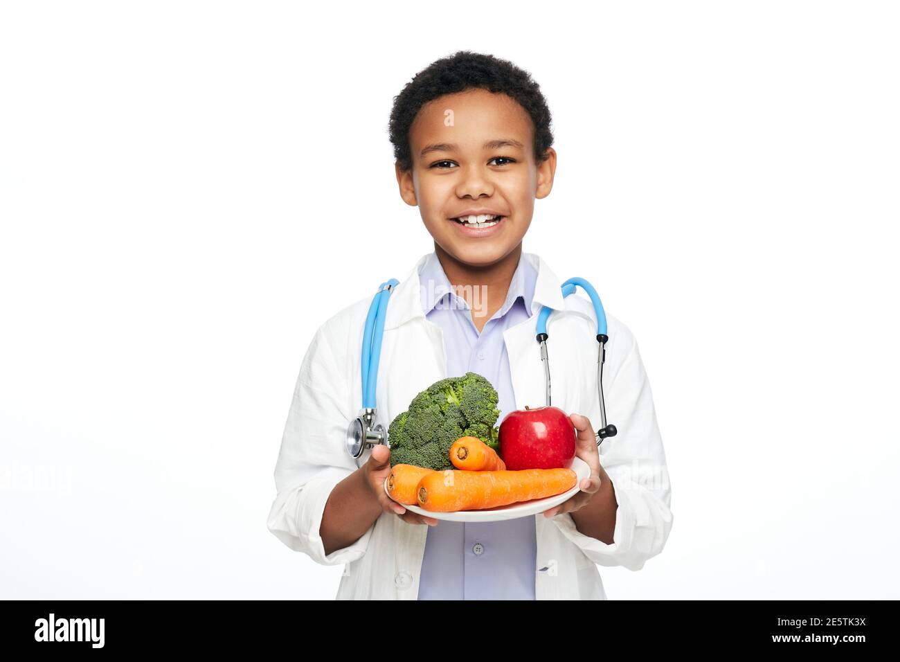 Junge afroamerikanische Kind, als Arzt gekleidet, empfiehlt Gemüse und Obst für die richtige Ernährung und Vorteile für Ihre Gesundheit Stockfoto