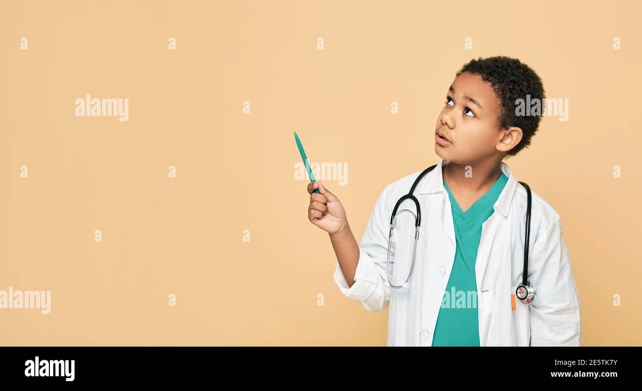 Serious African American Kind spielt Arzt, zeigt mit Stift auf leerem Raum auf beigem Hintergrund. Ort für Ihren Text Stockfoto