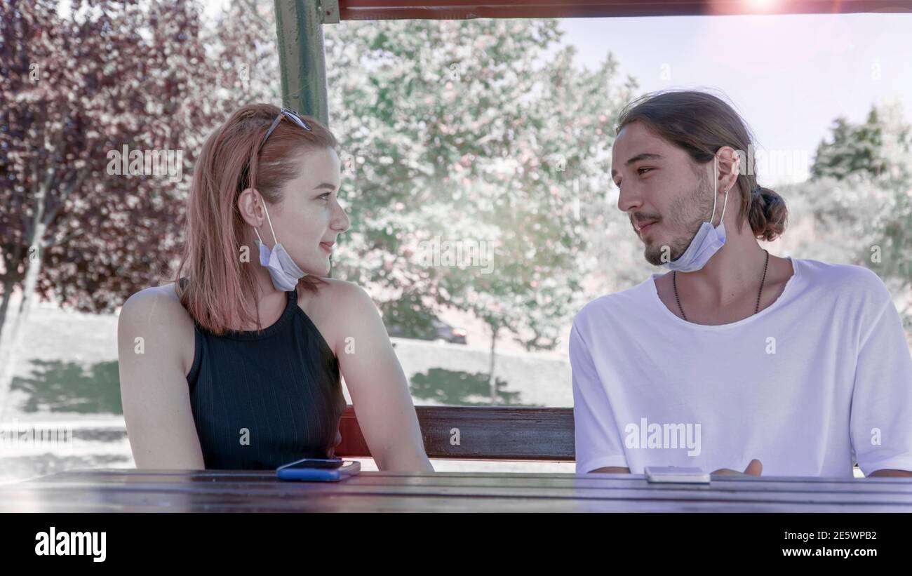 Junge Freunde tragen Gesichtsmasken und unterhalten sich, während sie auf einer Holzbank im Park sitzen. Freunde treffen und Spaß während der Pandemie haben Stockfoto