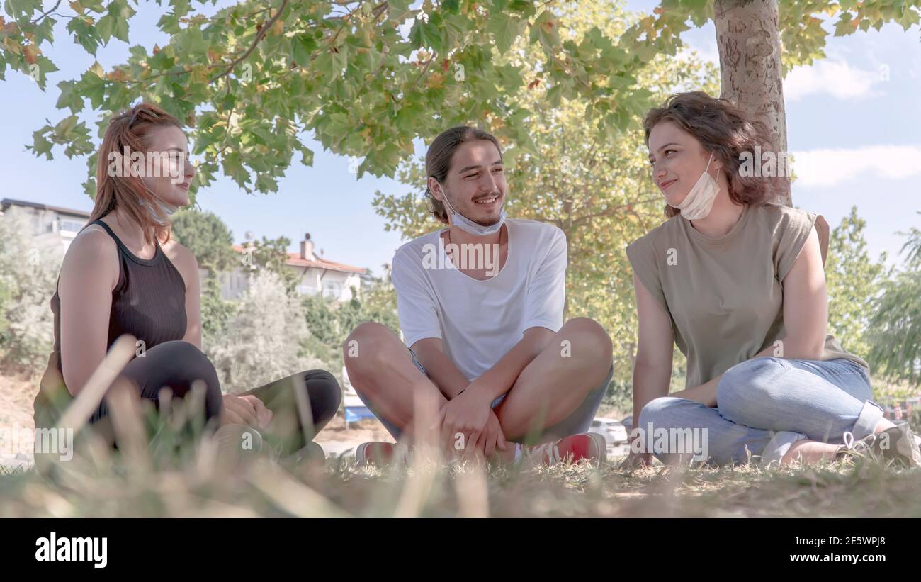 Junge Freunde reden und lachen, während sie auf Gras sitzen. Treffen mit Freunden im Park während der Coronavirus-Epidemie. Stockfoto