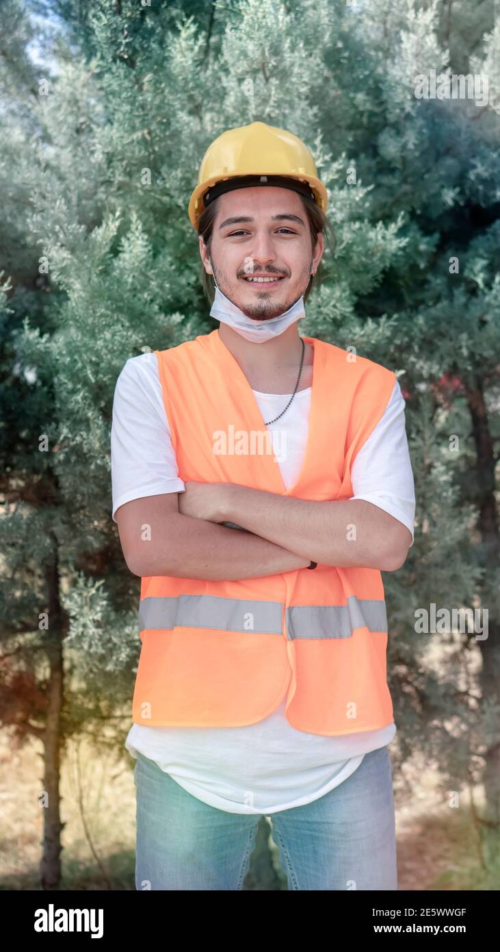 Portrait eines jungen Bauarbeiters mit gelbem Helm und Gesichtsmaske, der mit gekreuzten Armen steht und auf der Baustelle auf die Kamera schaut. Stockfoto