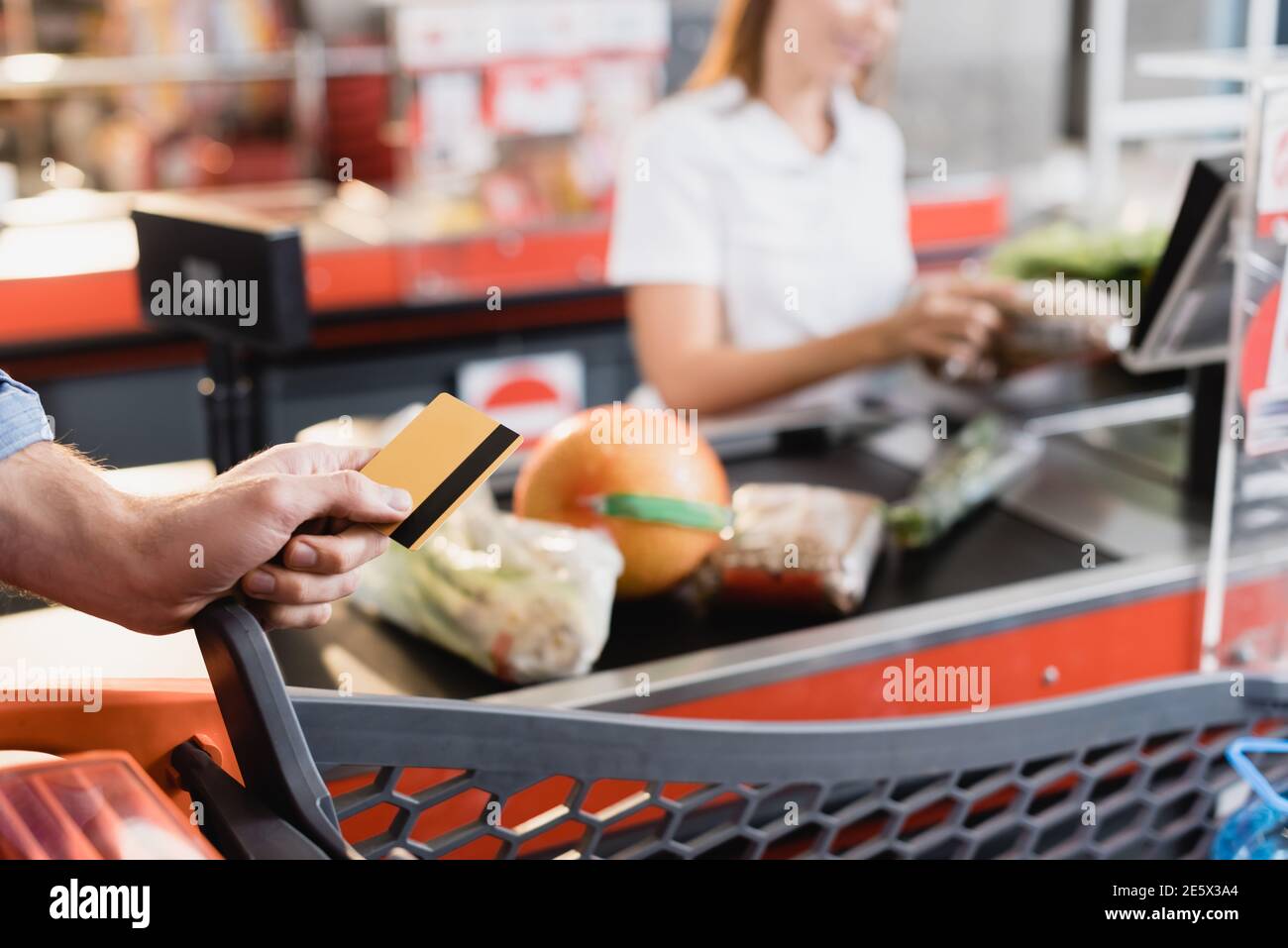 Beschnittene Ansicht des Mannes, der die Kreditkarte in der Nähe des Einkaufswagens hält Und Supermarkt Kasse auf verschwommenem Hintergrund Stockfoto