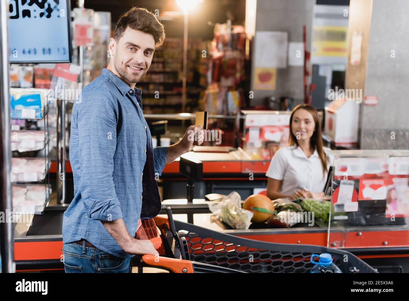 Lächelnder Mann mit Kreditkarte in der Nähe des Einkaufswagens und des Supermarkts Auschecken auf unscharfem Hintergrund Stockfoto