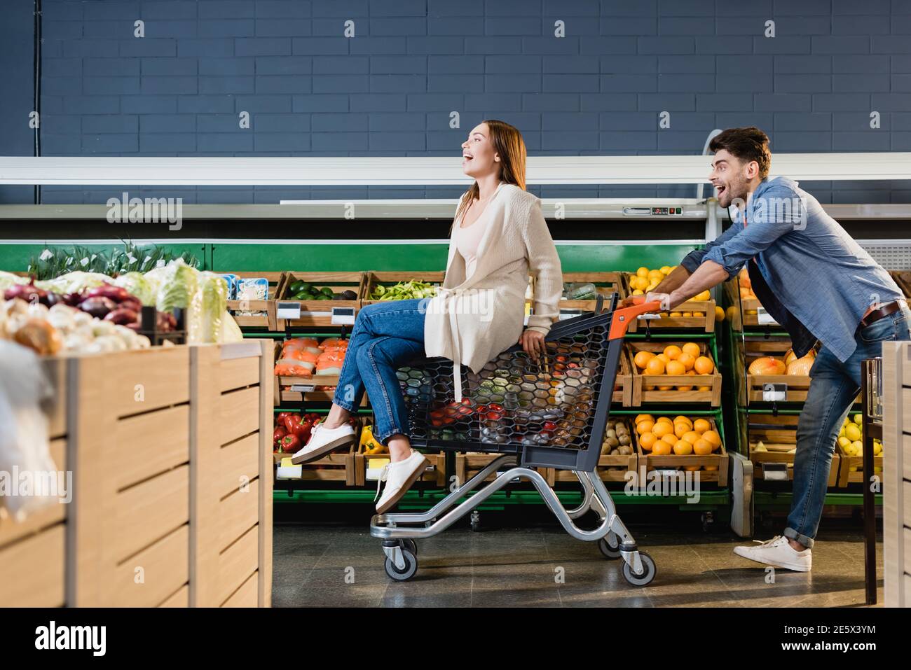 Fröhliches Paar rollt auf dem Einkaufswagen in der Nähe von Lebensmitteln im Supermarkt Stockfoto