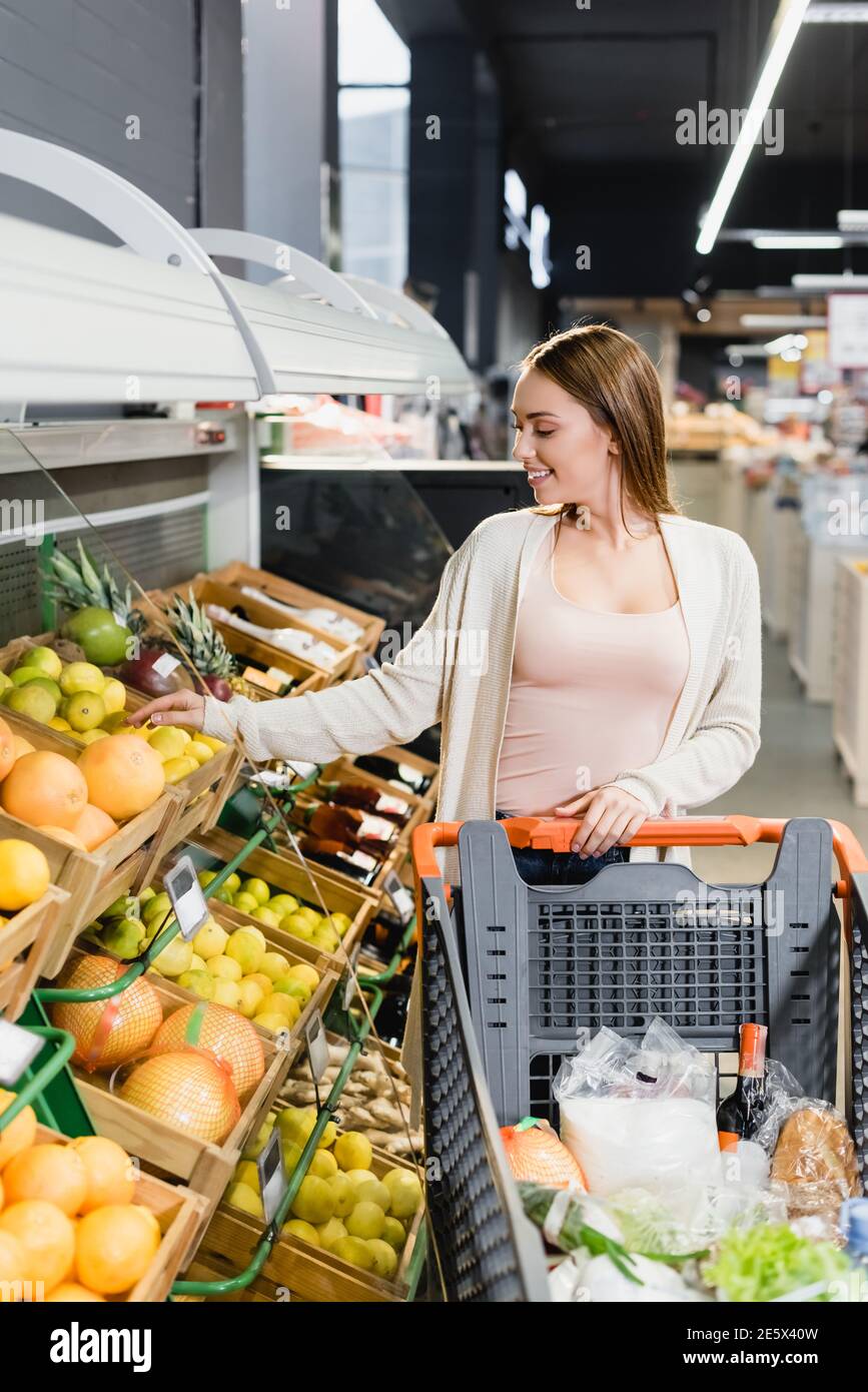 Fröhliche Frau, die Grapefruit in der Nähe von Früchten und Warenkorb in Supermarkt Stockfoto