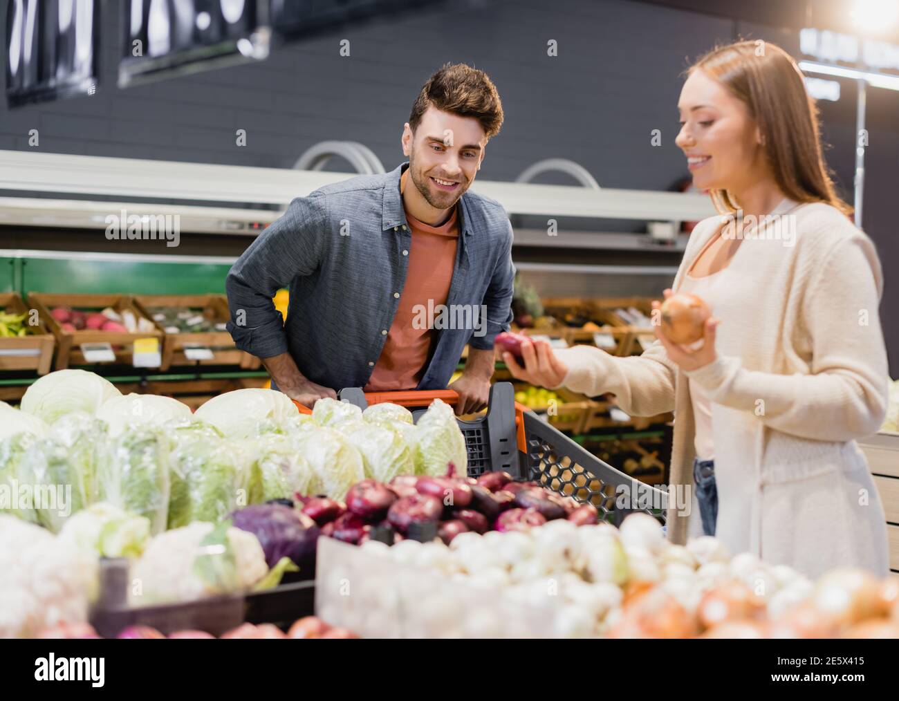 Lächelnder Mann, der in der Nähe des Einkaufswagens stand und Freundin mit Zwiebeln Auf verschwommenem Vordergrund im Supermarkt Stockfoto