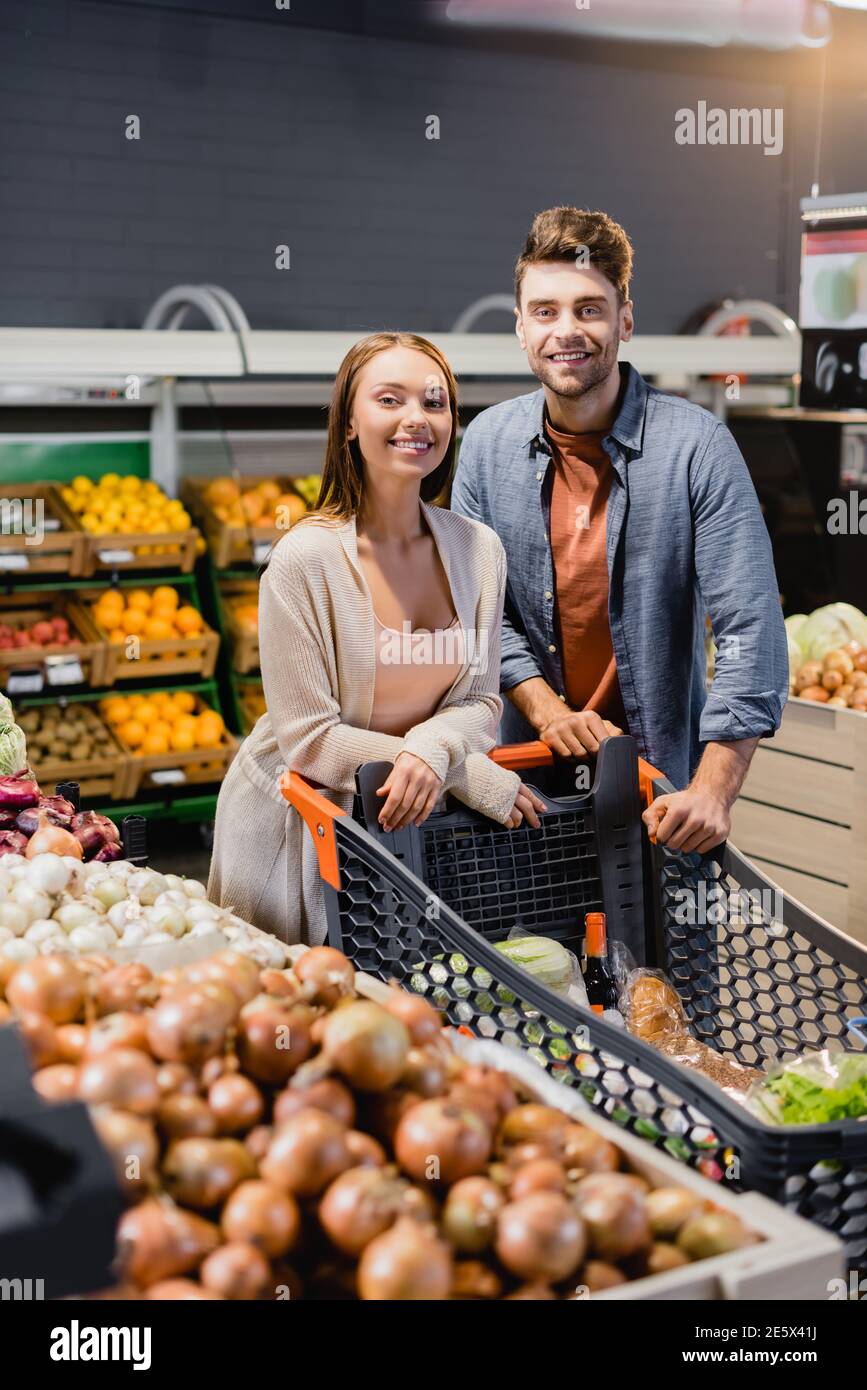 Lächelndes Paar, das in der Nähe des Einkaufswagens und des Gemüses im Supermarkt steht Stockfoto