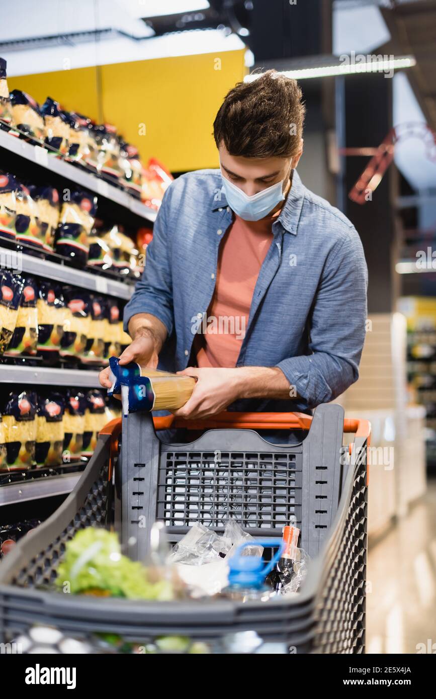 Mann in medizinischer Maske hält Pakete mit Lebensmitteln in der Nähe von Einkaufsmöglichkeiten Wagen auf verschwommenem Vordergrund im Supermarkt Stockfoto