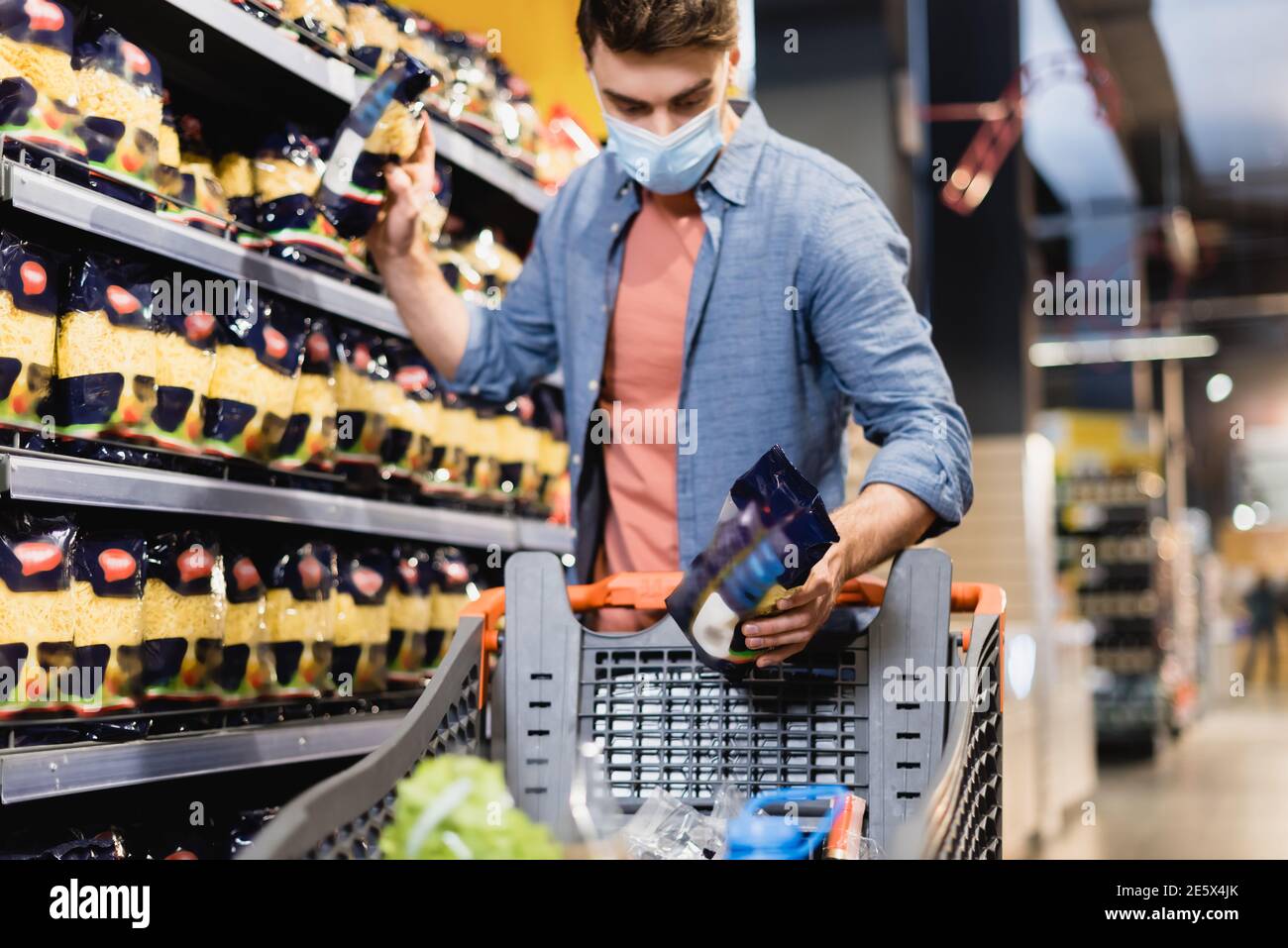 Mann in medizinischer Maske, der Lebensmittel in der Nähe von Regalen und Einkaufen nimmt Warenkorb im Supermarkt Stockfoto