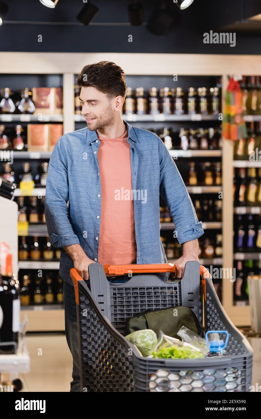 Lächelnder Mann, der mit dem Einkaufswagen auf verschwommenem Vordergrund läuft Supermarkt Stockfoto