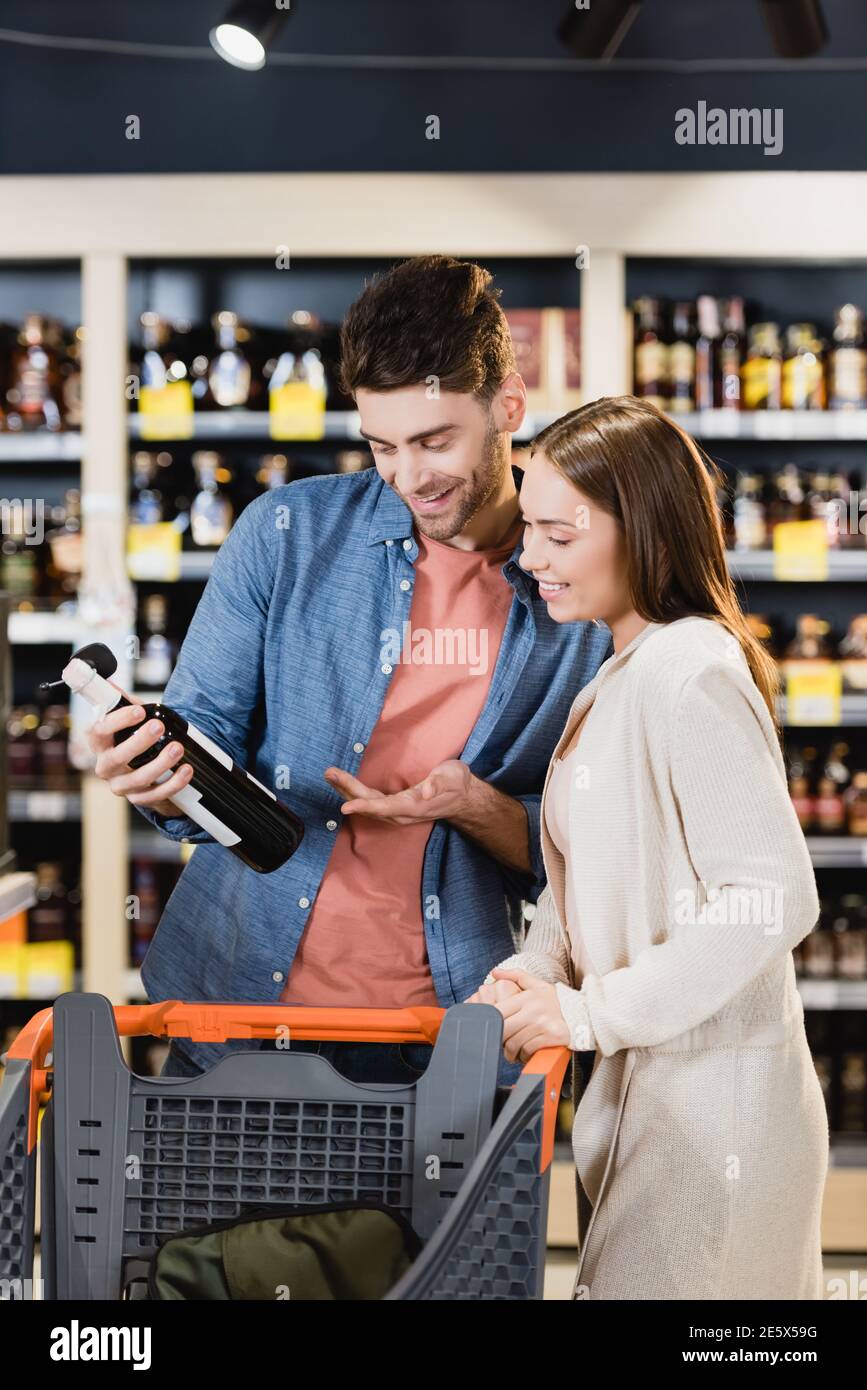 Fröhliches Paar, das in der Nähe des Einkaufswagens Weinflasche ansah Supermarkt Stockfoto