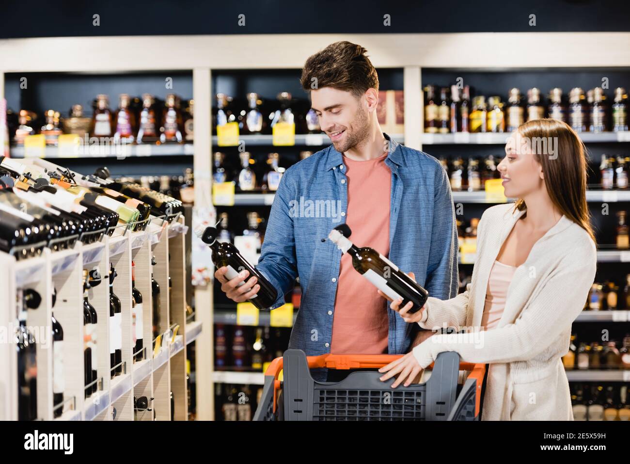 Ein junges Paar lächelt, während es in der Nähe des Einkaufsbuffens Weinflaschen hält Trolley im Supermarkt Stockfoto