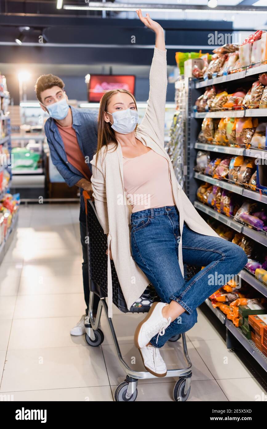Frau in medizinischer Maske sitzt auf Einkaufwagen in der Nähe von Freund Auf unscharfem Hintergrund im Supermarkt Stockfoto