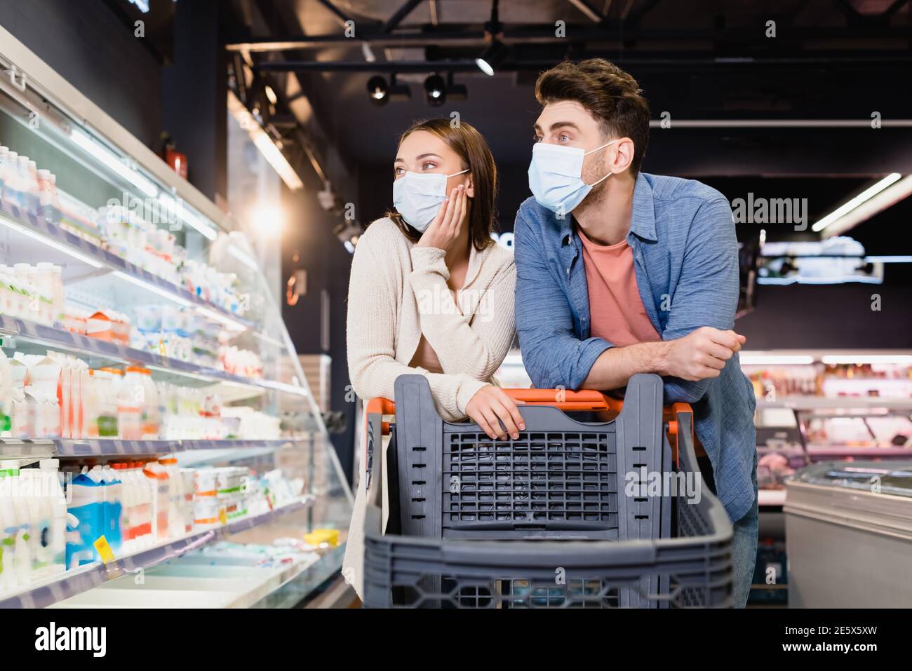 Paar in medizinischen Masken Blick auf Lebensmittel in der Nähe von Einkaufswagen Im Supermarkt Stockfoto