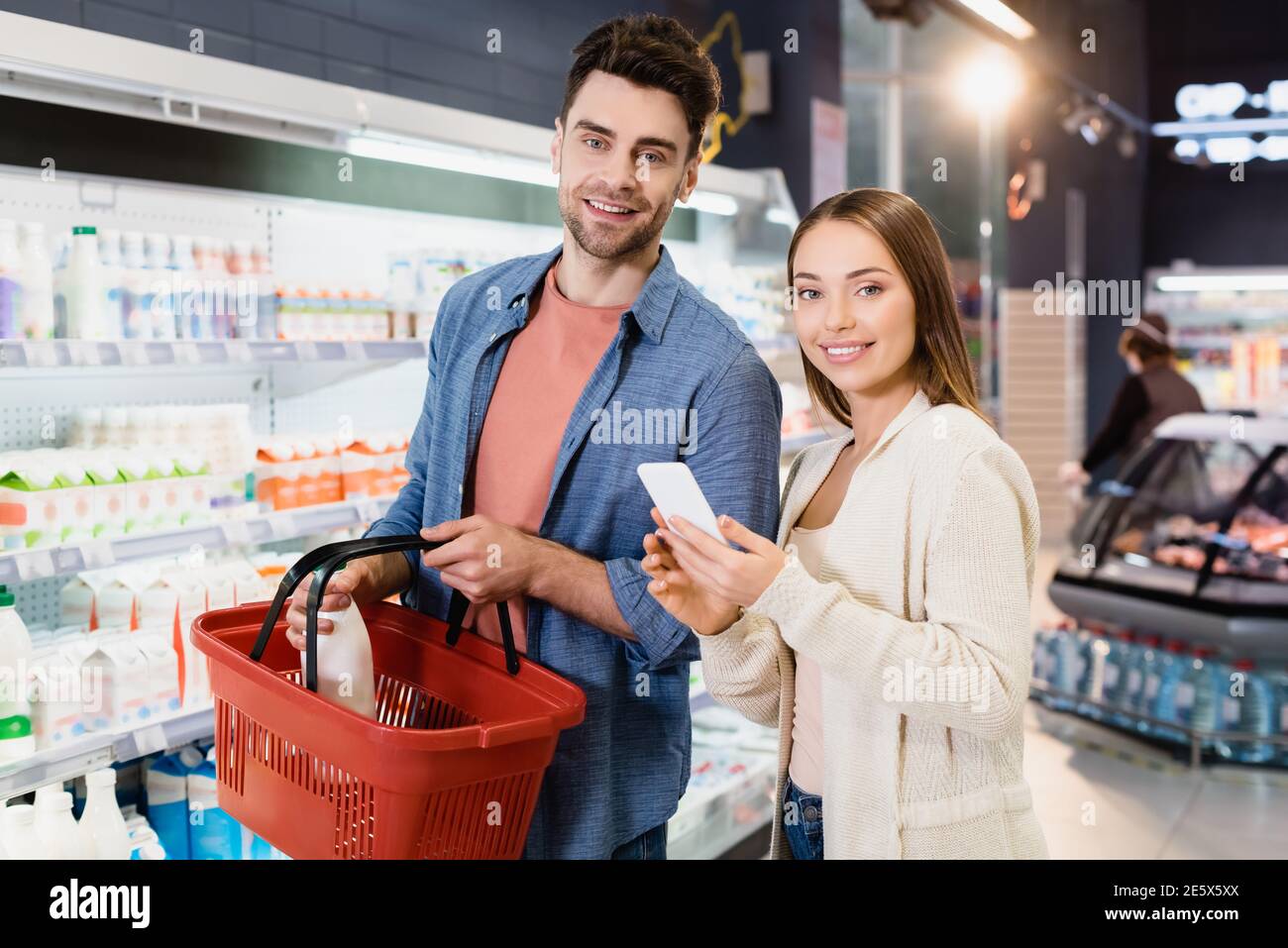 Lächelndes Paar mit Smartphone und Warenkorb Blick auf die Kamera Im Supermarkt Stockfoto