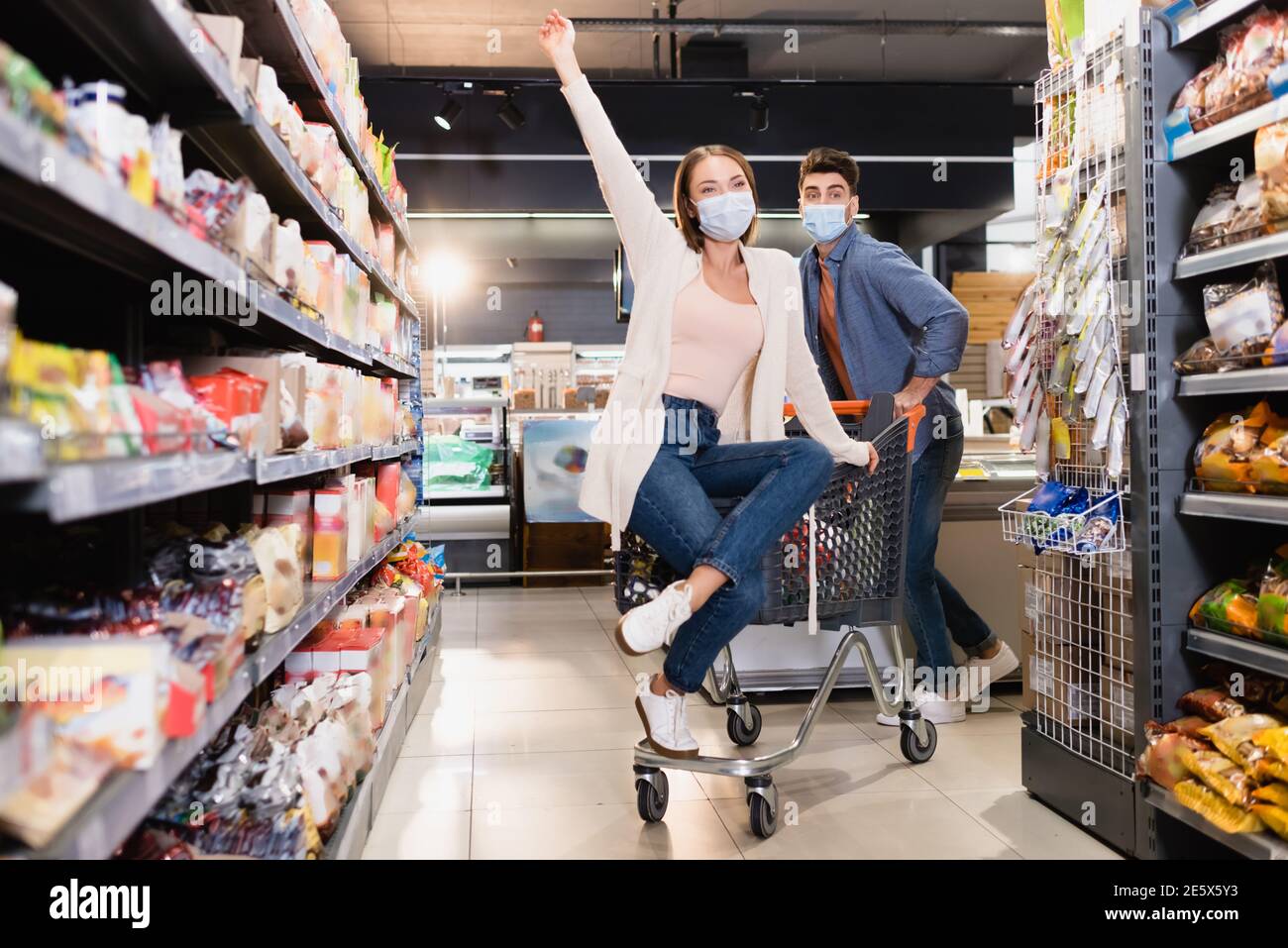Junge Frau in medizinischer Maske sitzt auf Shopping-Trolley in der Nähe Freund und Lebensmittel im Supermarkt Stockfoto