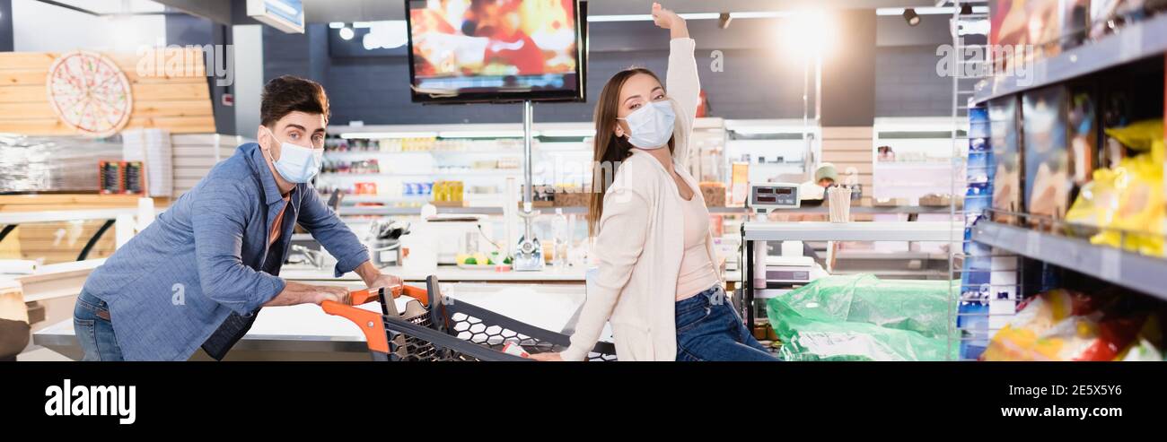 Mann in medizinischer Maske rollender Einkaufwagen mit Freundin im Supermarkt auf verschwommenem Vordergrund, Banner Stockfoto