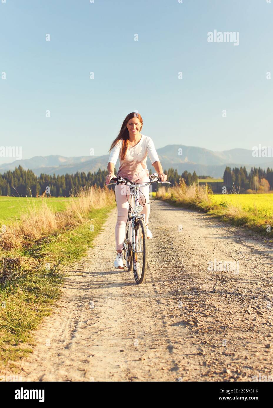 Junge Frau im Sommer leichte Kleidung fährt Fahrrad auf staubigen Straße in Richtung Kamera, Nachmittag Sonne scheint auf Felder und Wald im Hintergrund Stockfoto