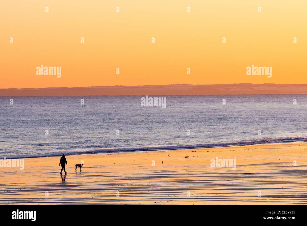 Eine einzige Person geht mit ihrem Hund an einem leeren Strand, wenn die Sonne untergeht, die warmen Farben des Himmels spiegeln sich im nassen Sand. Stockfoto