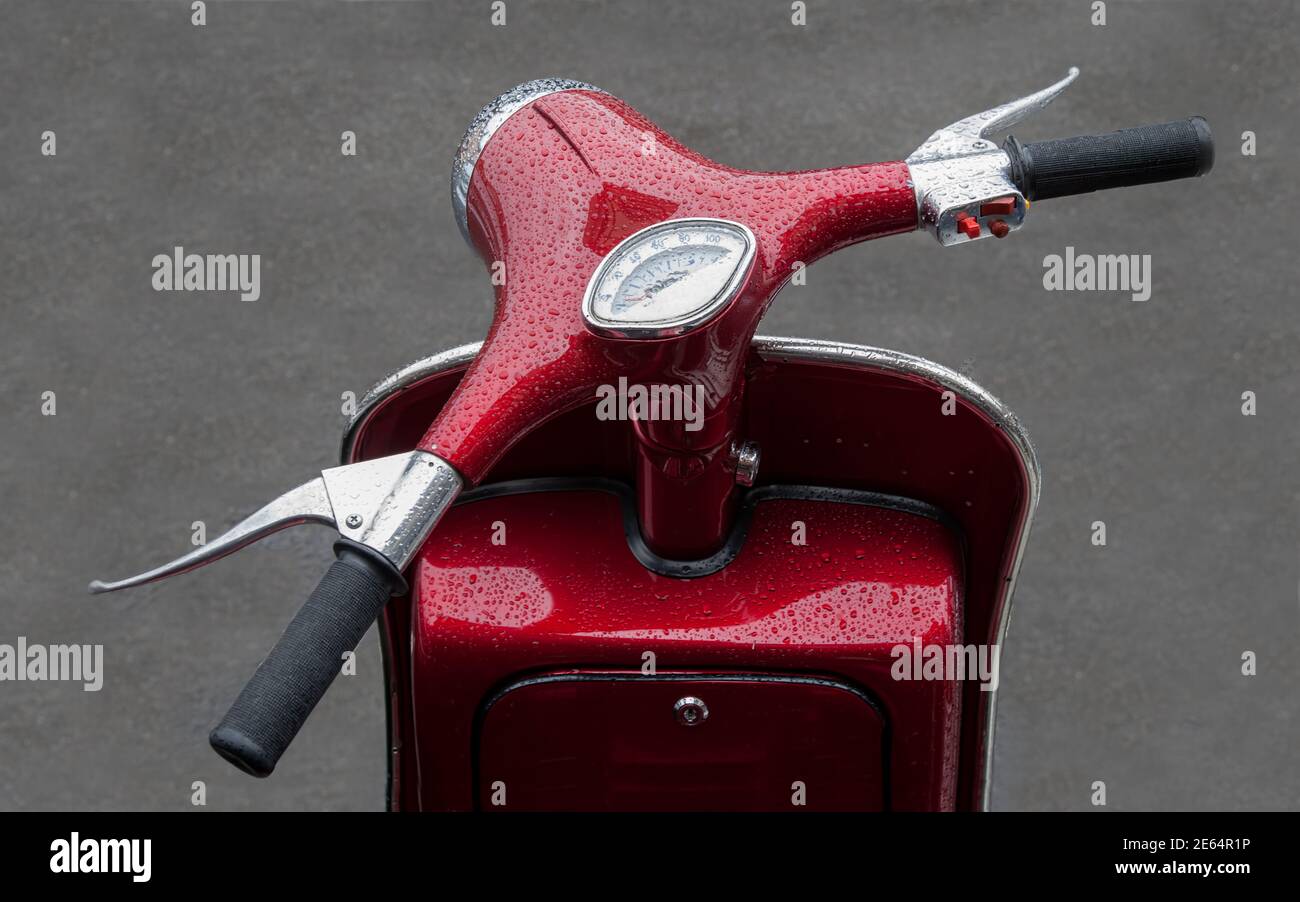 Eine Nahaufnahme eines roten Motorrollers zeigt nur die Front mit dem Lenker, Tachometer und Kontrollen. Die Lackierung hat Wassertropfen nach Regen Stockfoto
