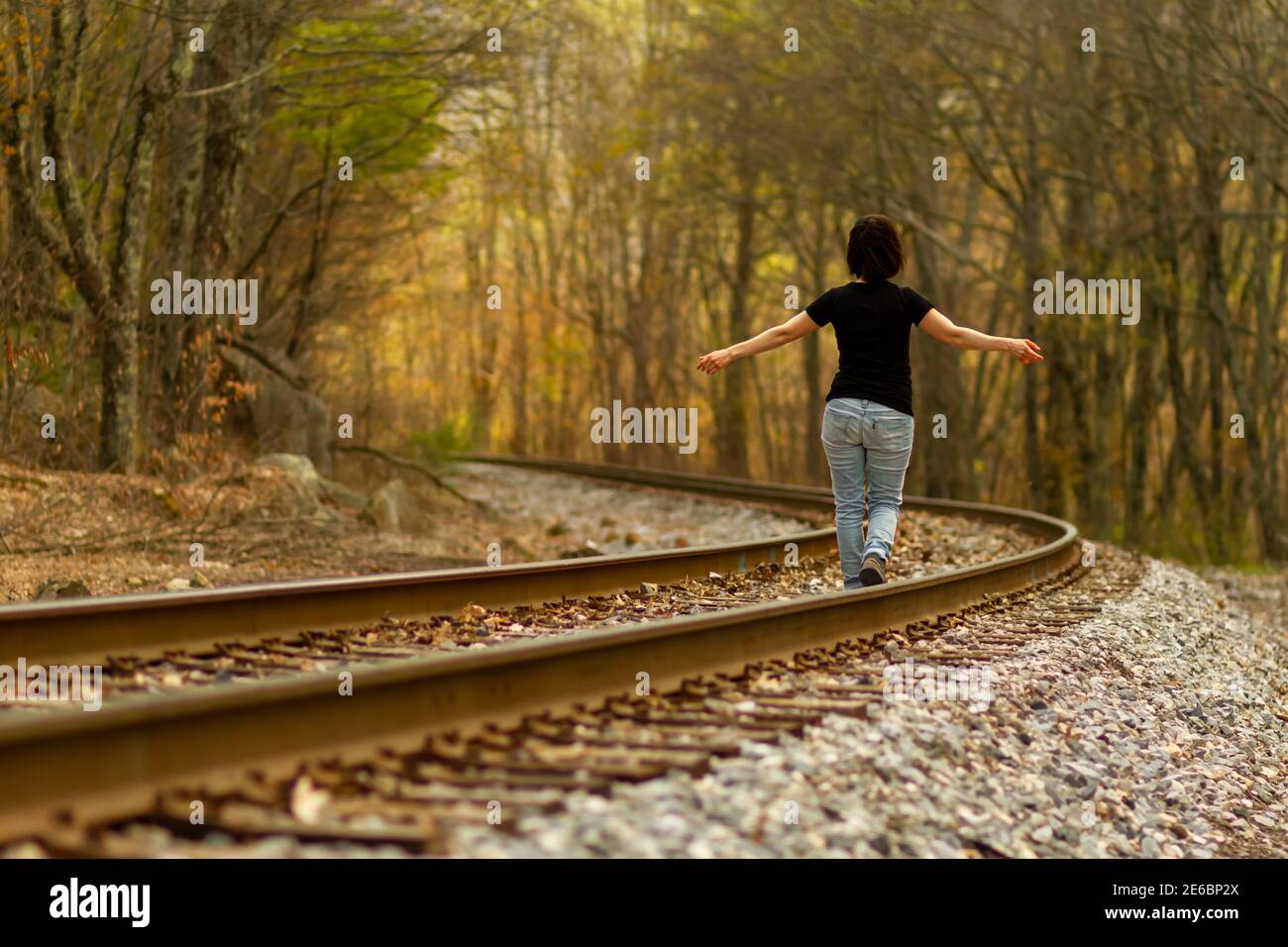Eine junge kaukasische Frau mit Jeans und T-Shirt geht mit weit geöffneten Armen auf den Bahngleisen an einem ländlichen Ort. Ein stimmungsvolle Konzept für f Stockfoto
