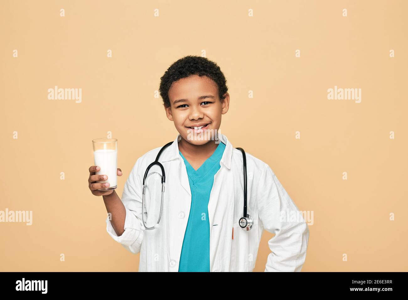 Afroamerikanischer Junge, gekleidet wie ein Arzt, hält ein Glas Milch auf beigem Hintergrund. Vorteile von Milch für Kinder Stockfoto