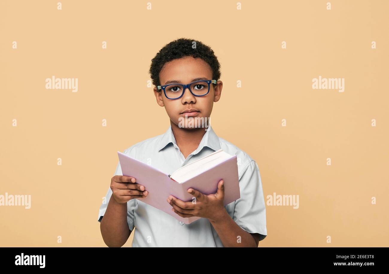 Serious afroamerikanischen kleinen Jungen trägt Brillen hält Buch Blick nachdenklich auf die Kamera, auf beigem Hintergrund Stockfoto