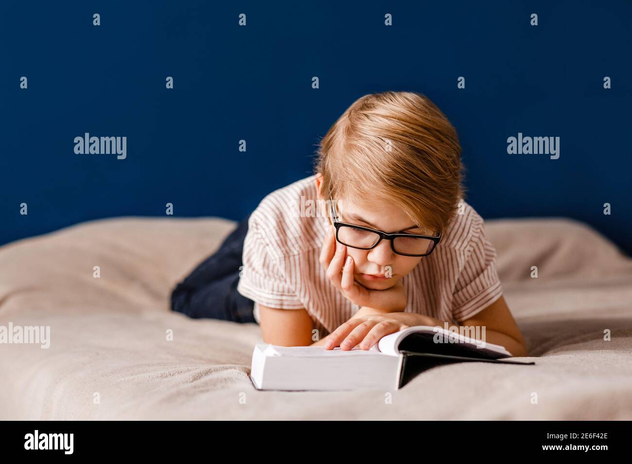 Ein 10-jähriger Junge mit Brille liegt auf dem Bett und liest ein großes Buch. Stockfoto