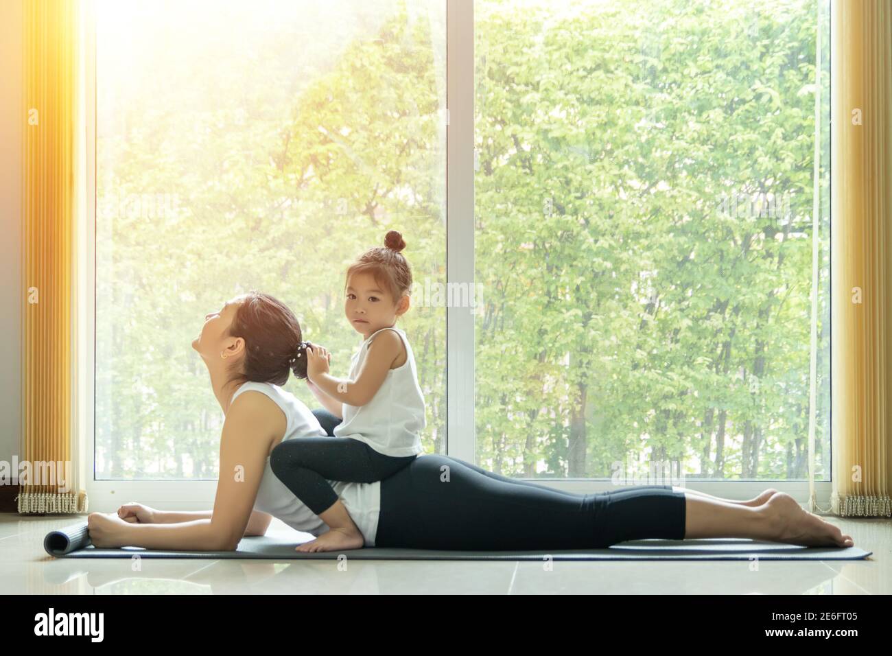Asiatische Mutter Praxis Cobra Yoga Pose, trainieren zu Hause mit einem entzückenden Tochter sitzen auf ihrem Rücken, Aktivität zu tun zusammen zu Hause in der Familie Stockfoto