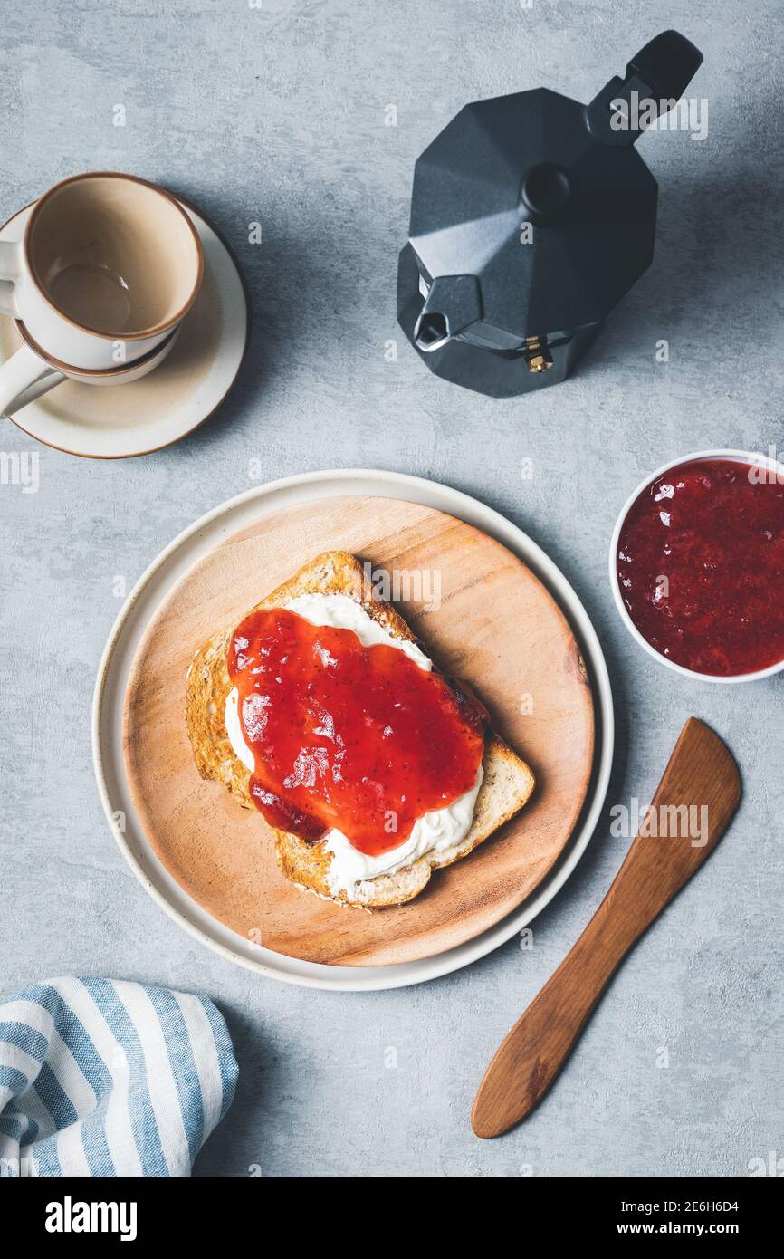 Layout der Frühstücksszene mit Toast, Frischkäse, Erdbeermarmelade und Kaffee auf einem grauen strukturierten Hintergrund. Stockfoto