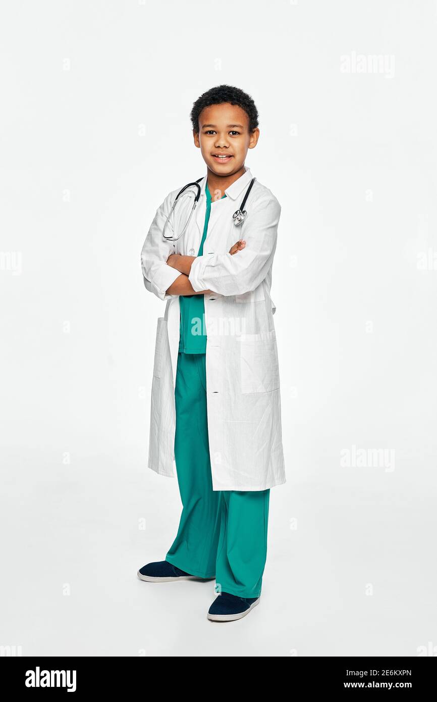 Lächelnder afroamerikanischer Junge, der einen Arztmantel mit gekreuzten Armen trägt, posiert und die Kamera in voller Länge anschaut Stockfoto