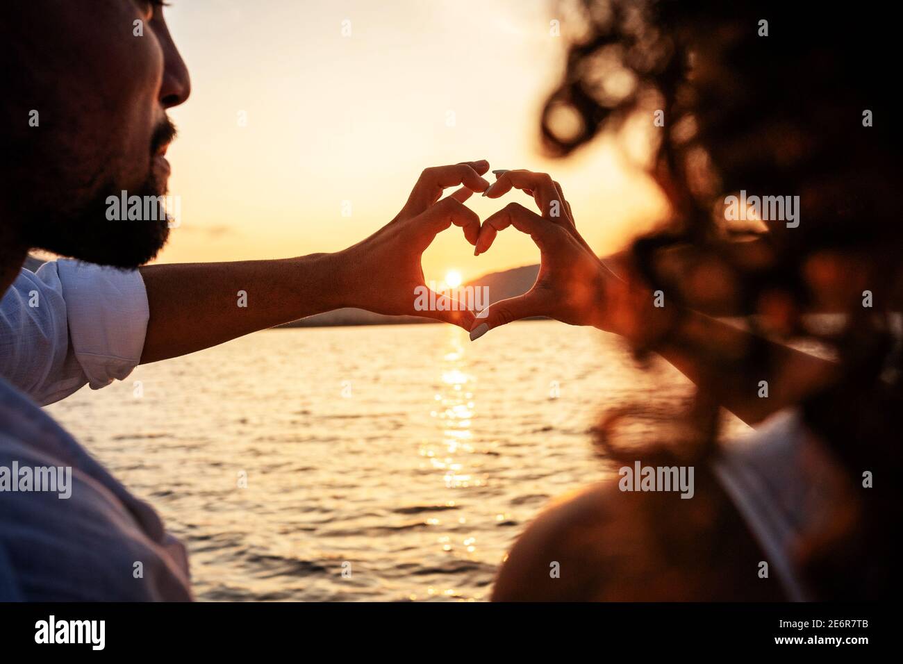 Blick von hinten auf ein junges, schönes Paar, das mit den Fingern Herzform  macht und auf den Sonnenuntergang hinter den Bergen blickt, der das Licht  im Meer reflektiert Stockfotografie - Alamy
