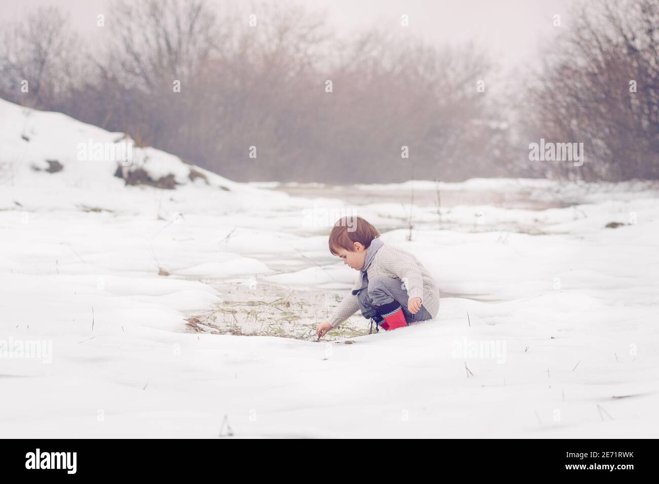 Kleines kaukasisches Kind in Schal gewickelt trägt einen Pullover, spielen im Winterschnee und Pfütze. Stockfoto