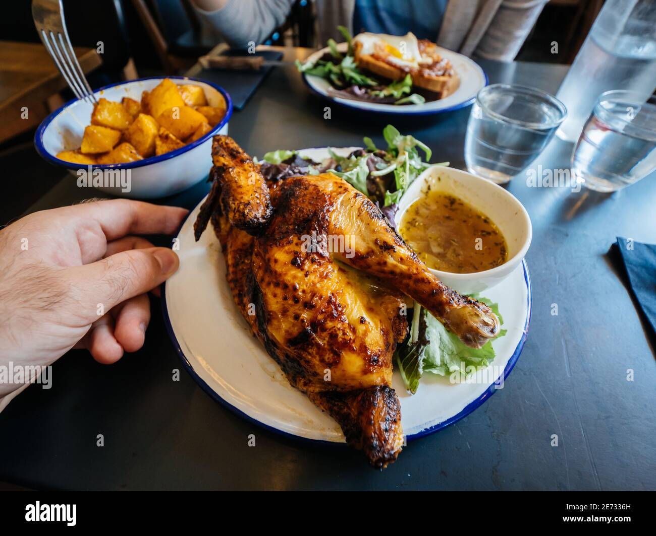 POV Persona perspektivische Sicht männliche Hand Halteplatte Mit leckeren gebratenen halben Huhn auf einem Restauranttisch mit Bratkartoffeln und leckere Bouillon Stockfoto