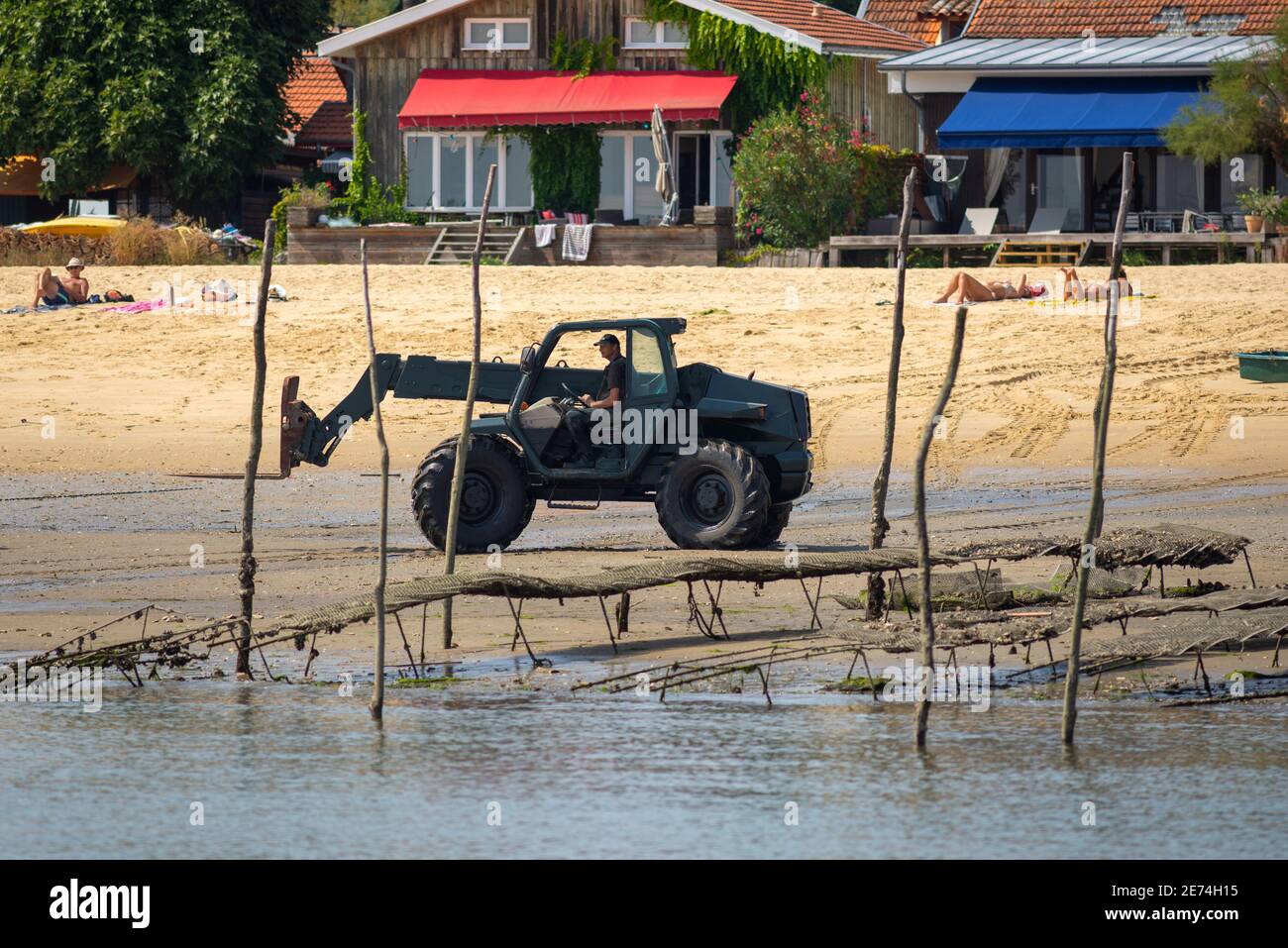 Ein Traktor arbeitet am Strand bei Ebbe in der Nähe von Cap Ferret. Austernfalle am Strand traditionelle Holzhäuser im Hintergrund Stockfoto