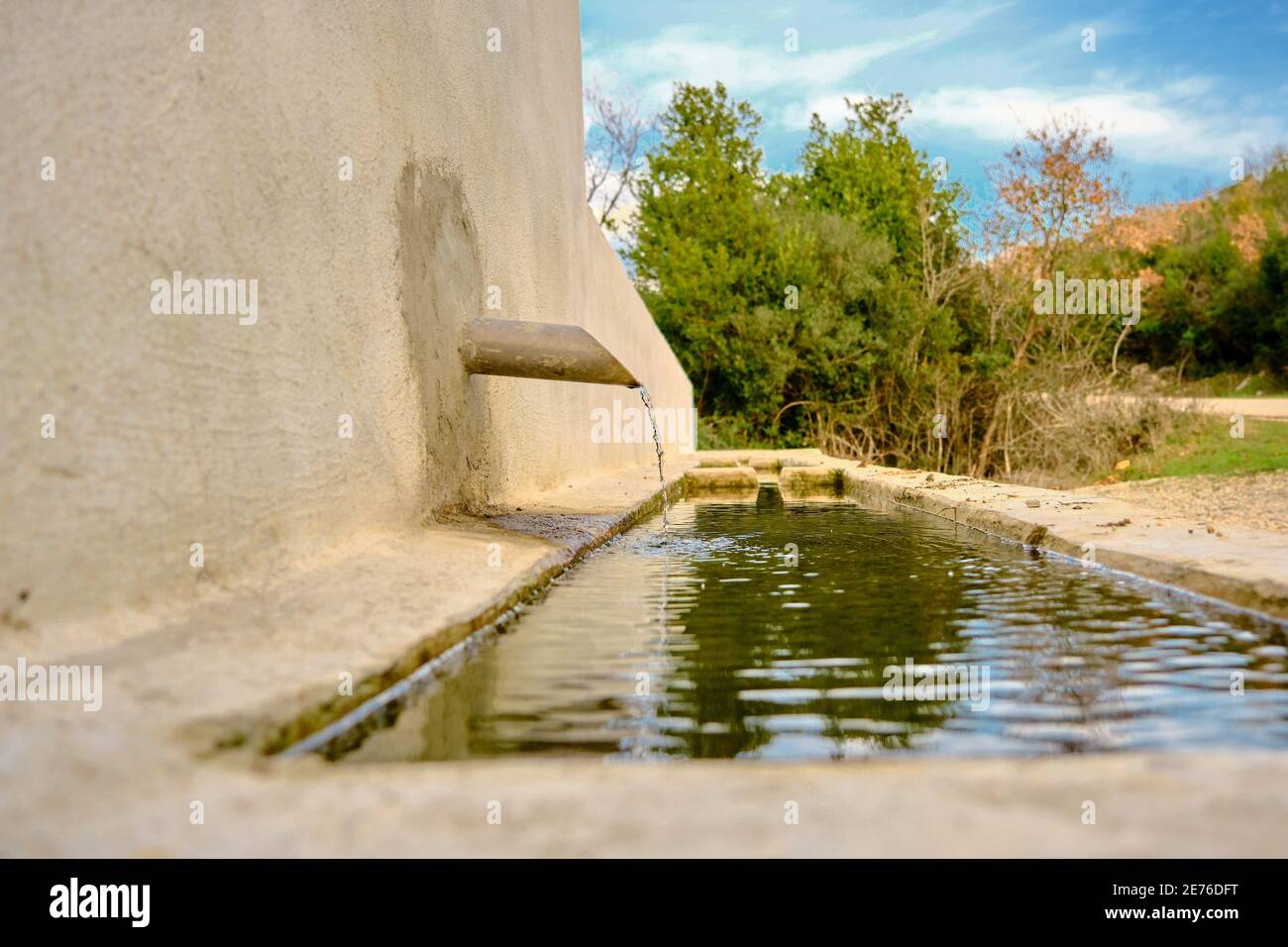 Quelle und natürlich trinkbares Wasser und Brunnen draußen in der Nähe der Schotterstraße in der Landschaft. Kleiner Hügel, viele Arten von Pflanzen und wolkigen Himmel Hintergrund Stockfoto