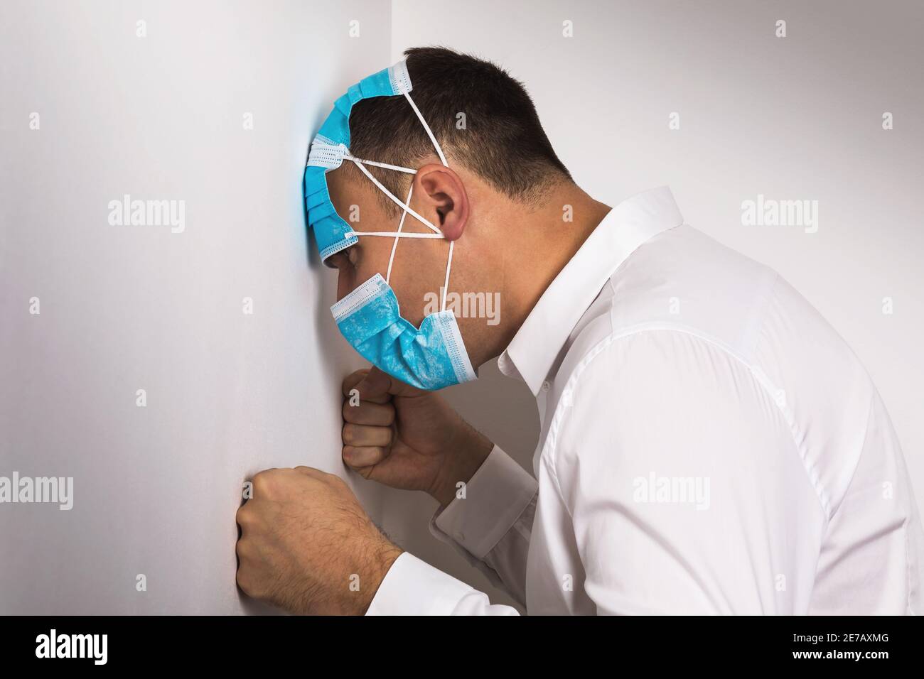 Ein Mann in mehreren medizinischen Masken gekleidet, der seinen Kopf und seine Hände gegen die Wand lehnte. Das Konzept der Depression aufgrund der Quarantäne durch die COVI verursacht Stockfoto