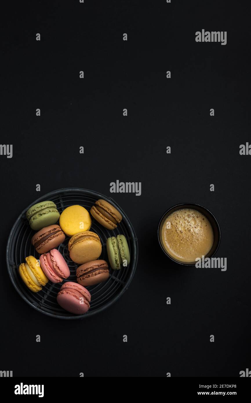 Horizontale und vertikale hausgemachte Makronen in einem eingekreisten Drahttablett neben einer Kaffeetasse auf schwarzem Hintergrund. Stockfoto