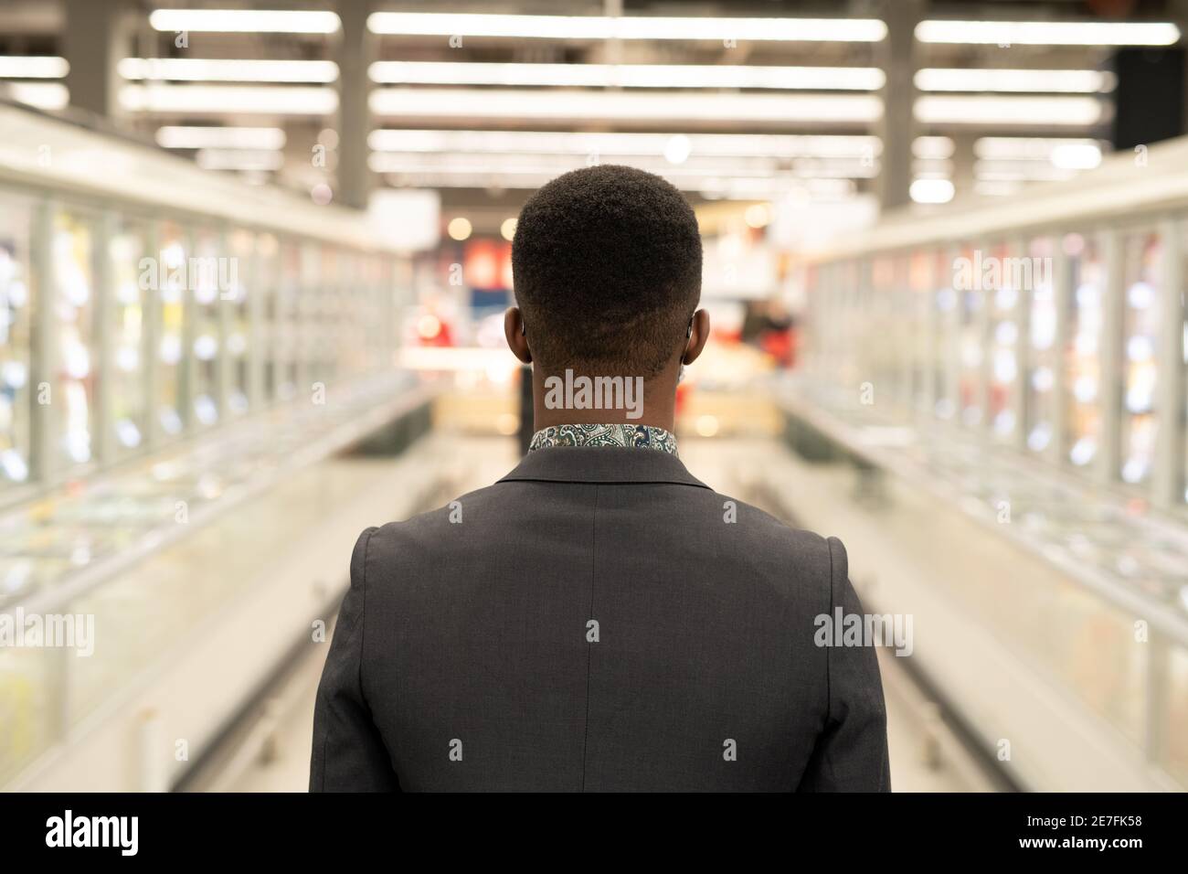 Rückansicht des jungen eleganten afrikanischen Geschäftsmann bewegt sich zwischen großen Und lange Displays mit Tiefkühlkost im modernen Supermarkt Stockfoto