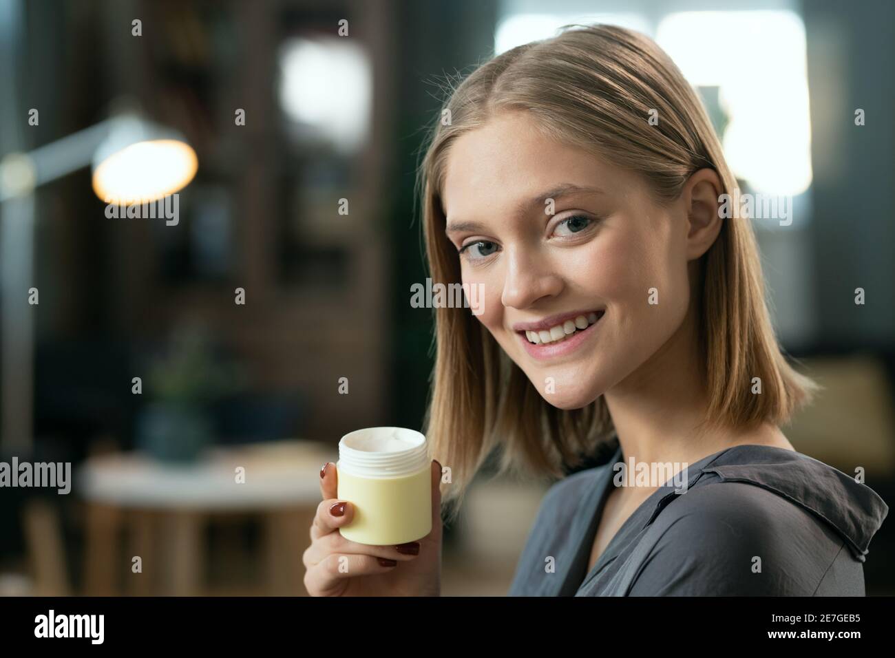 Junge hübsche Frau mit blonden Haaren hält kleine Glas mit Natürliche handgemachte kosmetische Produkt und Blick auf Sie mit toothy Lächeln Stockfoto