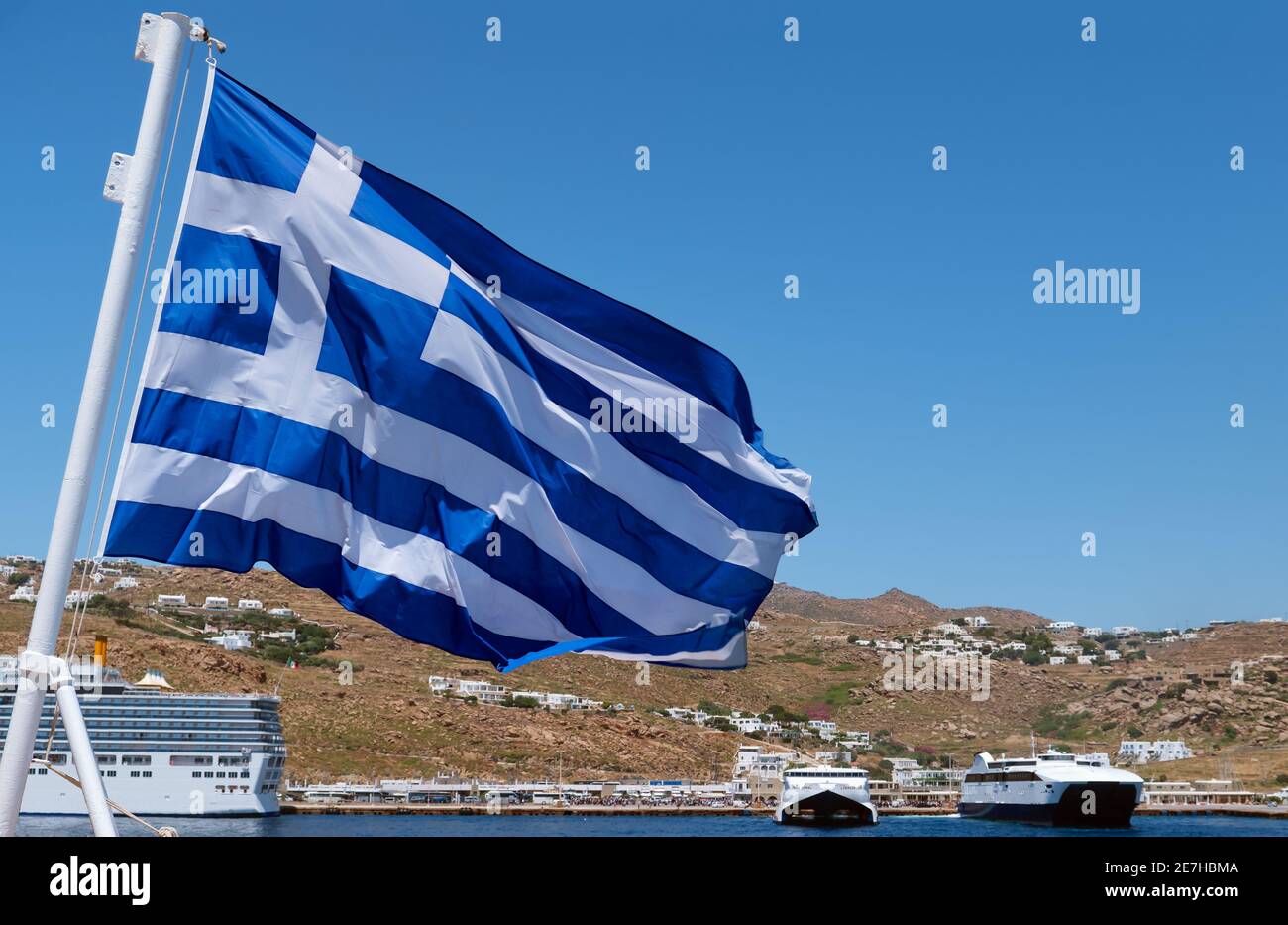 Griechische Flagge winkt gegen die typische griechische Insellandschaft. Kreuzfahrtschiffe und Fähren, Sommertag. Inselhüpfen, Abenteuer, Mittelmeer erkunden. Stockfoto