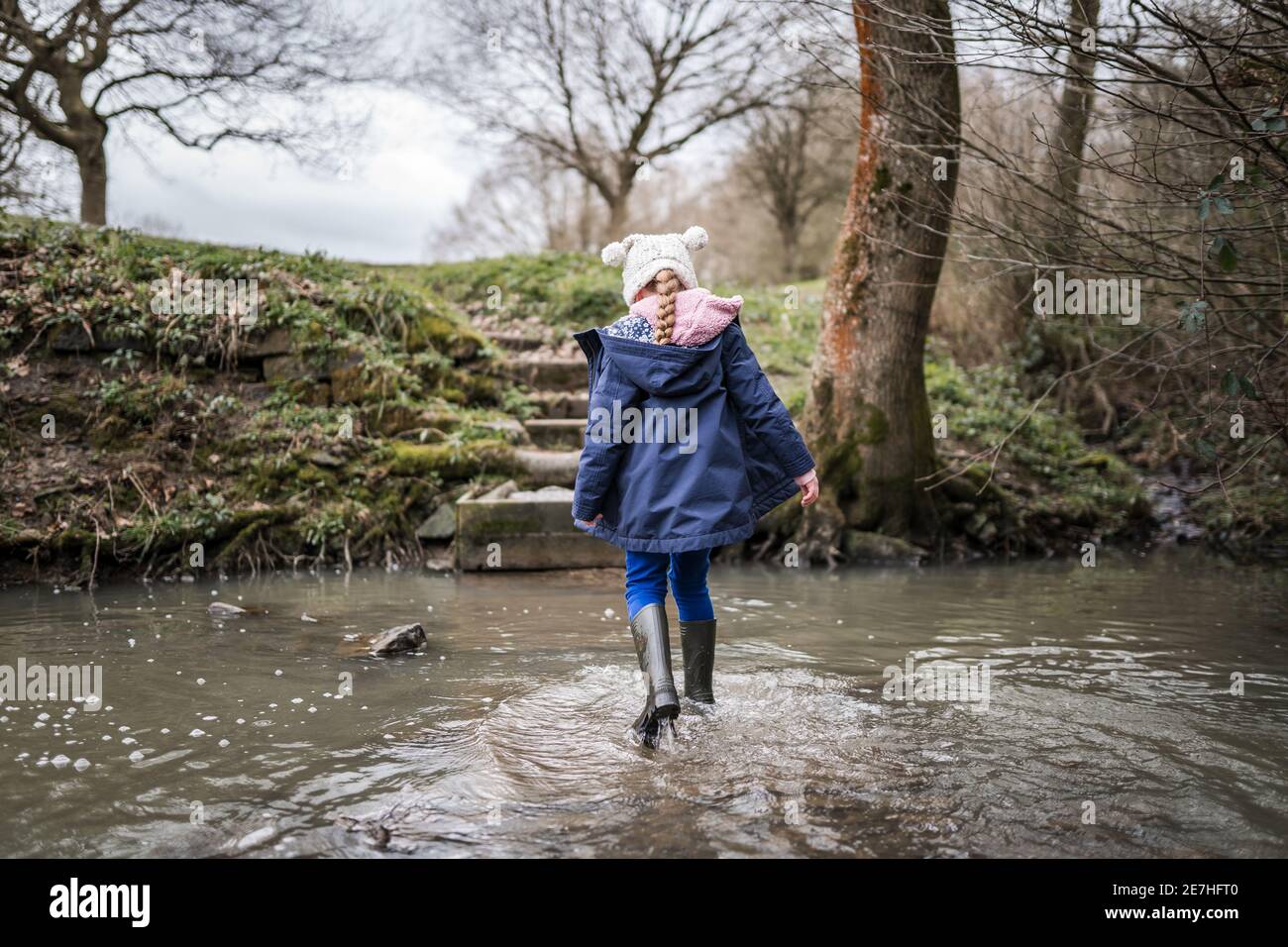 Aufgeregt glücklich junge Mädchen wellinton Stiefel zu Fuß durch die herbstliche Landschaft Flusslauf plantschen im Wasser Erforschen der freien Natur bobble hut Stockfoto