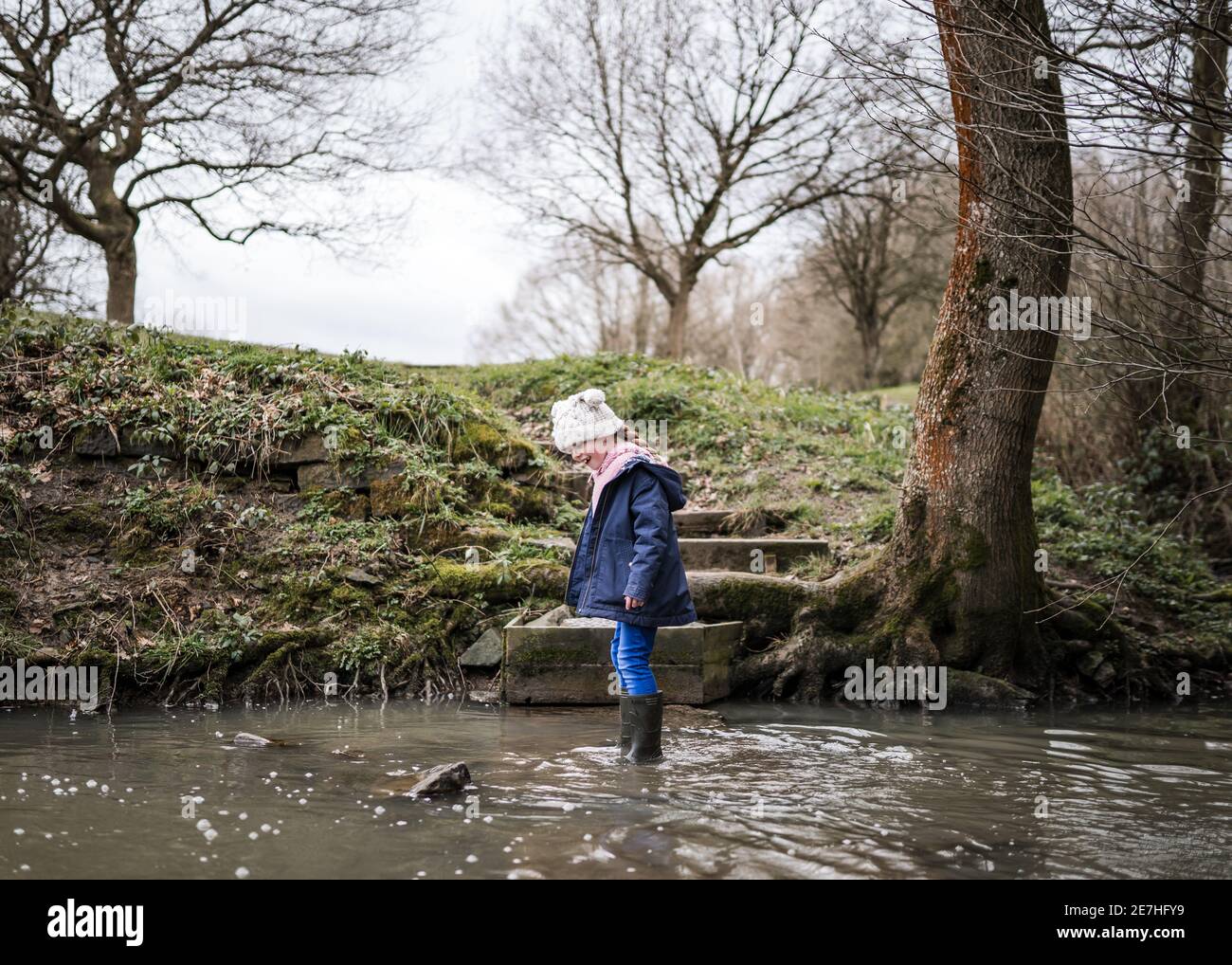 Aufgeregt glücklich junge Mädchen wellinton Stiefel zu Fuß durch die herbstliche Landschaft Flusslauf plantschen im Wasser Erforschen der freien Natur bobble hut Stockfoto
