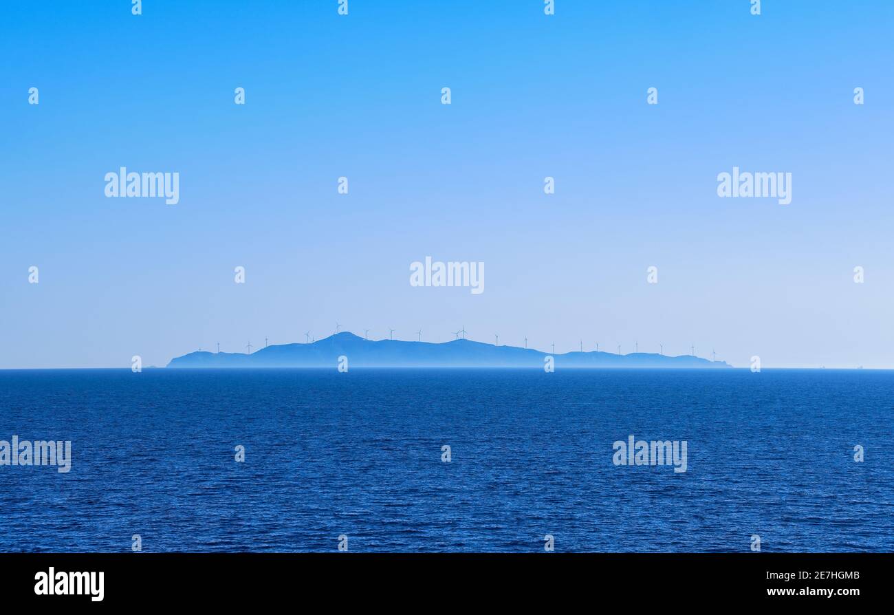 Meereslandschaft von ruhigen azurblauen Meer, blauen Himmel, keine Wolken. Entfernte Insel im Dunst mit Silhouetten von elektrischen Turbinen Windmühlen. Mittelmeer, Griechenland. Stockfoto