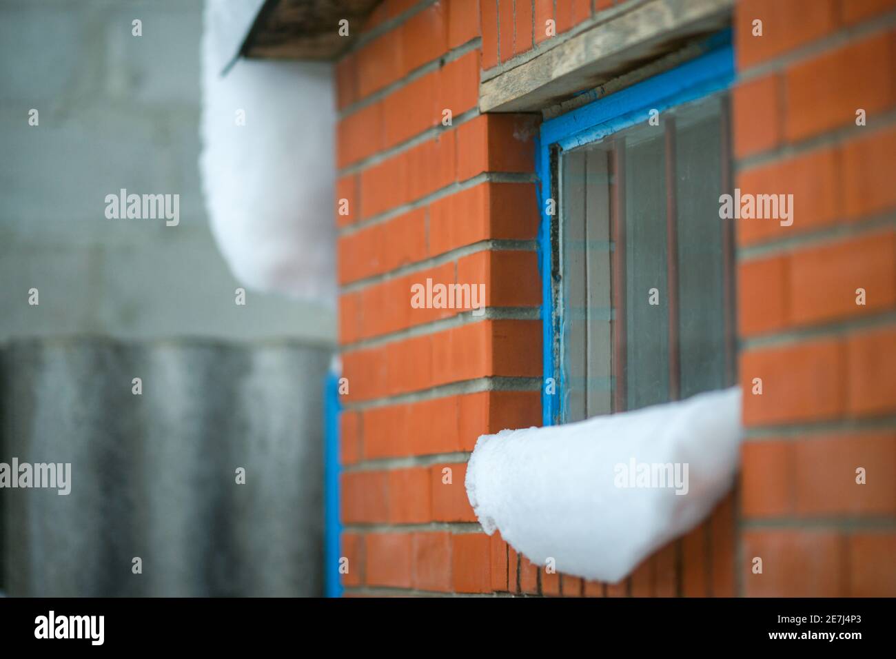 Schneekappen auf der Fensterbank. Eine Wand mit roten Ziegeln und einem blauen Fensterrahmen mit Balken. Stockfoto