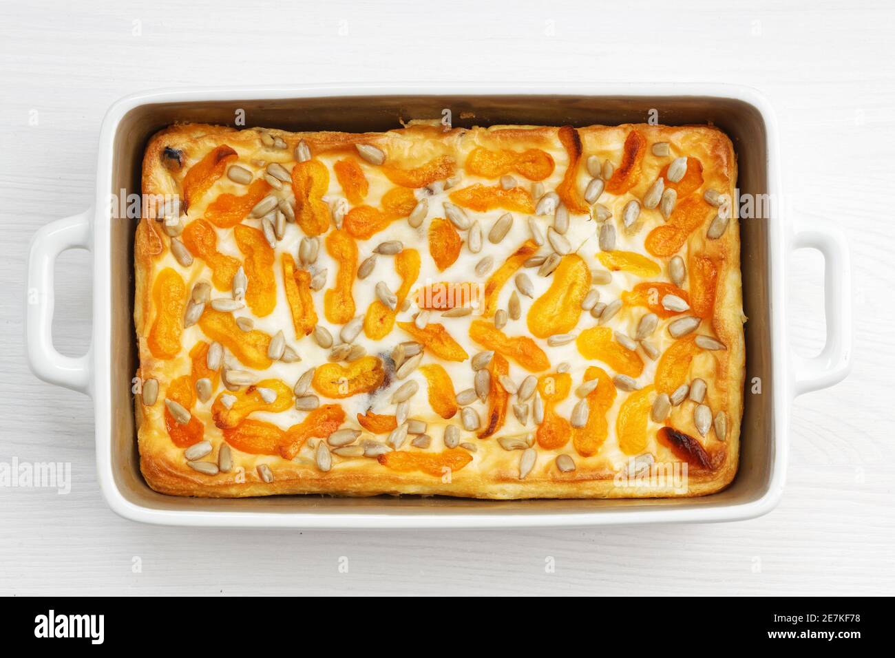 Hausgemachter Hüttenkäse-Auflauf mit getrockneten Aprikosen und Sonnenblumenkernen auf weißem Holztisch. Draufsicht. Stockfoto
