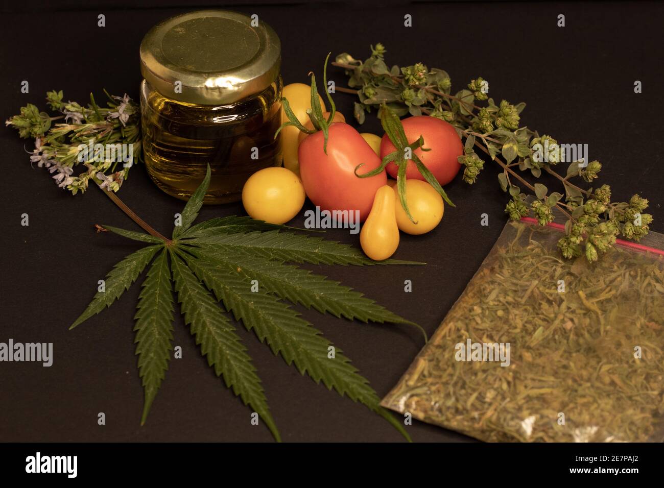 Ein Cannabisblatt, ein Glas mit cbd-Öl, Tomaten und aromatischen Kräutern auf schwarzem Hintergrund. Das Bild zeigt die Beziehung zwischen Nahrung und Cannabis Stockfoto