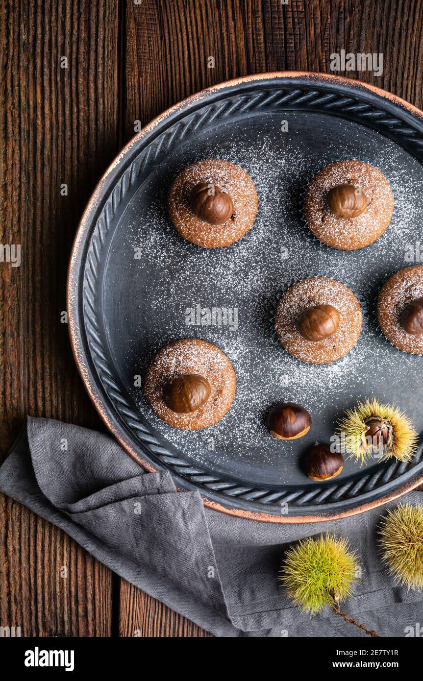 Muffins aus Edelkastanienpüree und Kakao, gekrönt mit geschälten und gebackenen Kastanien, bestäubt mit Puderzucker auf rustikalem Holzhintergrund Stockfoto